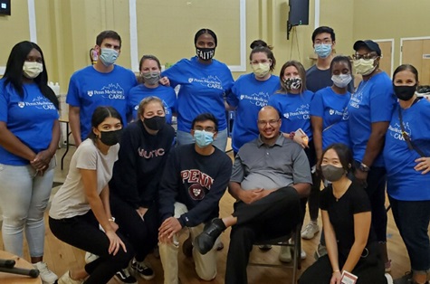 Nine Penn Medicine staff members wearing blue “Penn Medicine CARES” shirts pose together with other volunteers from the University of Pennsylvania. Nurse Amber Roseman is standing in the middle wearing a black and white mask that says “FAITH”. 